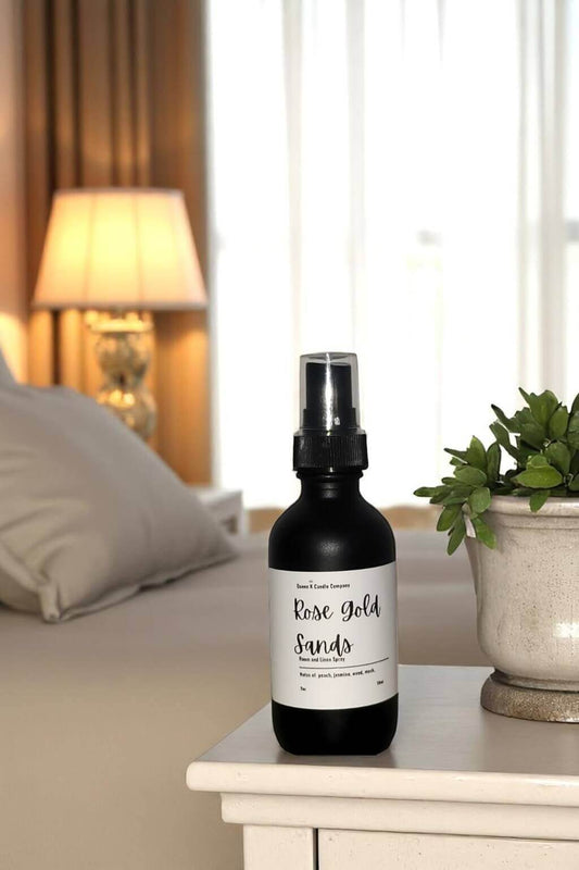 Black spray bottle labeled 'Rose Gold Sands' on a nightstand with a blurred bedroom background