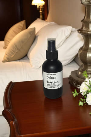 Black spray bottle labeled 'Deluxe Bourbon' on a wooden nightstand with pillows and a lamp in the background.