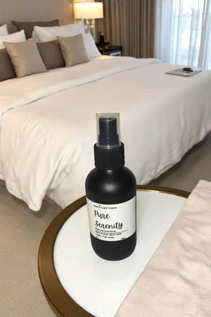 Black spray bottle labeled 'Pure Serenity' on a nightstand with a bed in the background