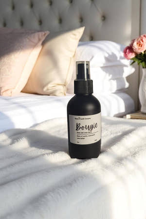 Black spray bottle labeled 'Bougie' on a bed with pillows and flowers.