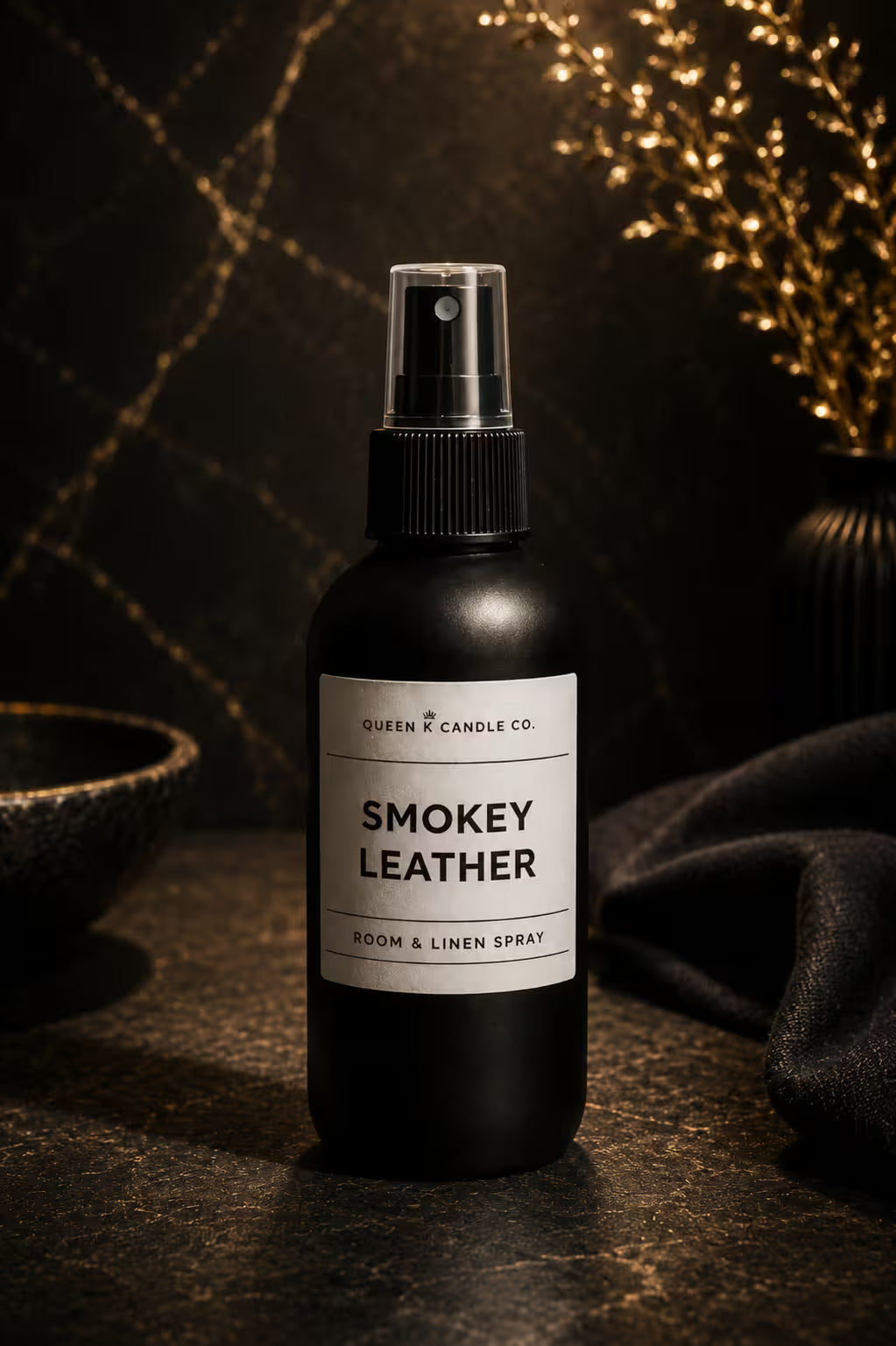Black room & linen spray bottle labeled 'Smokey Leather' by Queen & Candle Co. on a dark surface with decorative elements.